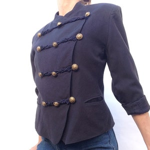 Pode incluir: Um blazer azul escuro com bot&otilde;es dourados e detalhes pretos. O blazer tem um colarinho mandarim e um fecho de um s&oacute; bot&atilde;o.