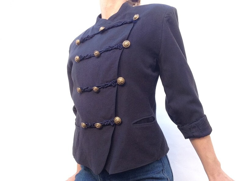 Pode incluir: Um blazer azul escuro com bot&otilde;es dourados e detalhes pretos. O blazer tem um colarinho mandarim e um fecho de um s&oacute; bot&atilde;o.