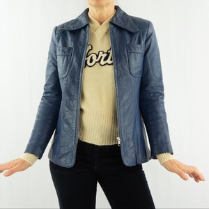 K&ouml;nnte beinhalten: Eine marineblaue Lederjacke mit Kragen und zwei Vordertaschen. Die Jacke wird &uuml;ber einem beigen Pullover mit dem Wort "forts" in schwarzer Schrift getragen. Das Model tr&auml;gt auch eine schwarze Hose.