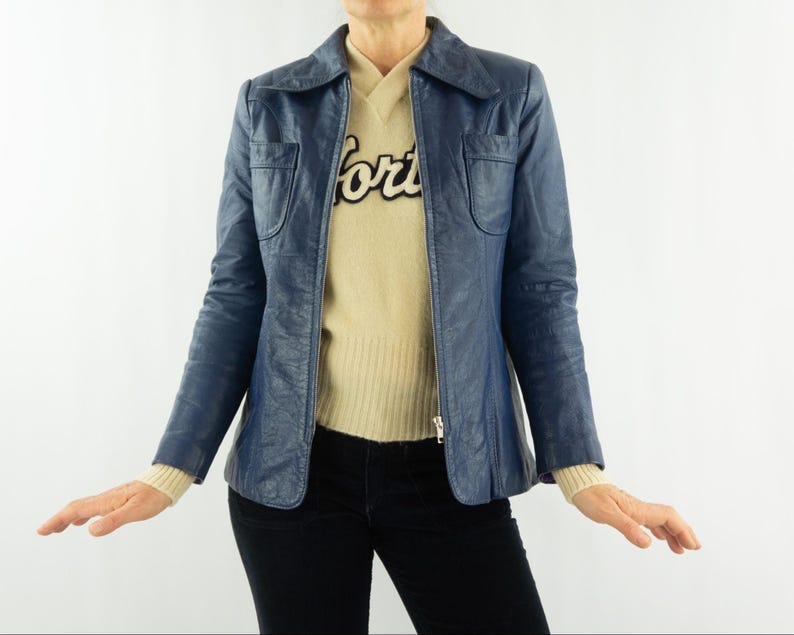 K&ouml;nnte beinhalten: Eine marineblaue Lederjacke mit Kragen und zwei Vordertaschen. Die Jacke wird &uuml;ber einem beigen Pullover mit dem Wort "forts" in schwarzer Schrift getragen. Das Model tr&auml;gt auch eine schwarze Hose.