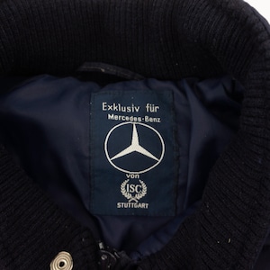 May include: A navy blue jacket with a white and blue striped detail on the sleeve. The jacket has a label that reads "Exklusiv f&uuml;r Mercedes-Benz von ISC STUTTGART" and features the Mercedes-Benz logo.