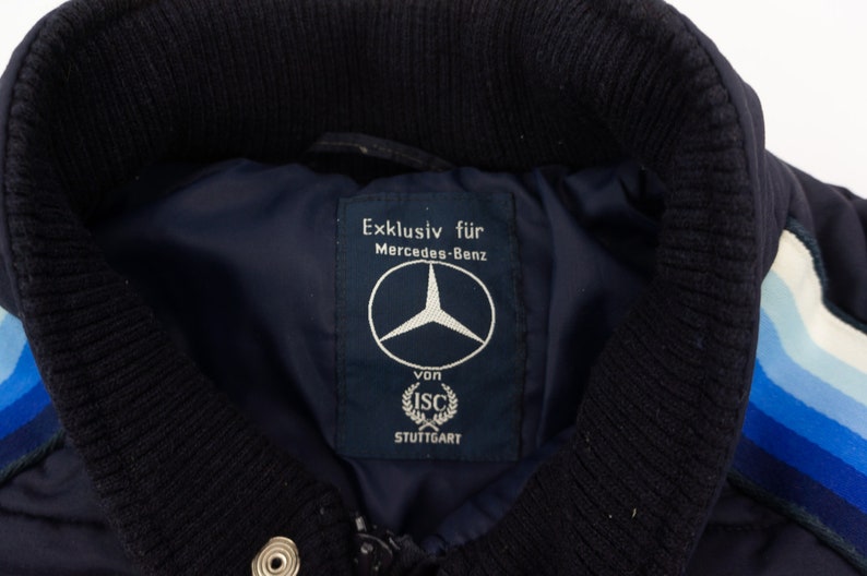 May include: A navy blue jacket with a white and blue striped detail on the sleeve. The jacket has a label that reads "Exklusiv f&uuml;r Mercedes-Benz von ISC STUTTGART" and features the Mercedes-Benz logo.