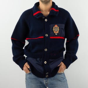 Puede incluir: Un cárdigan de punto azul marino con rayas rojas y un parche con un diseño de corona y rueda dorada. El cárdigan tiene cierre de botones y cuello.