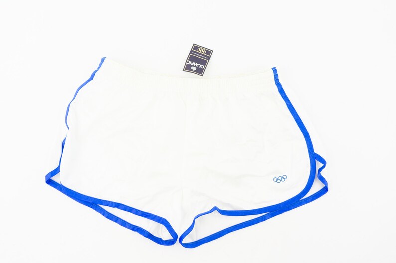 May include: White athletic shorts with blue trim and the Olympic rings logo. The shorts have a drawstring waist and are made of a lightweight fabric.