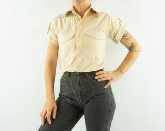 May include: A short-sleeved beige button-down shirt featuring a classic collar, two chest pockets with flaps, and rolled cuffs, paired with high-waisted dark charcoal gray denim jeans that have a visible button fly closure.