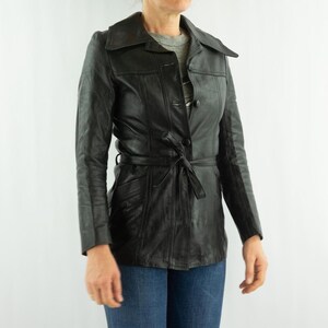 May include: Black leather jacket with a tie belt, button closure, and a collared neckline. The jacket has long sleeves and pockets. The person is wearing blue jeans.