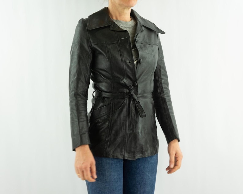 May include: Black leather jacket with a tie belt, button closure, and a collared neckline. The jacket has long sleeves and pockets. The person is wearing blue jeans.