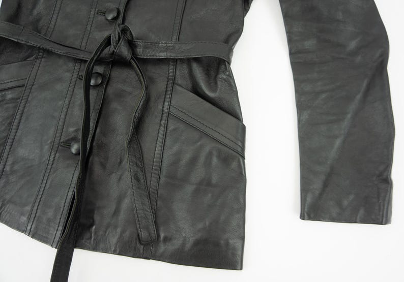 May include: Black leather jacket with a belted waist, button closures, and a pocket. The jacket has long sleeves and a tailored fit. The leather appears to be slightly distressed.