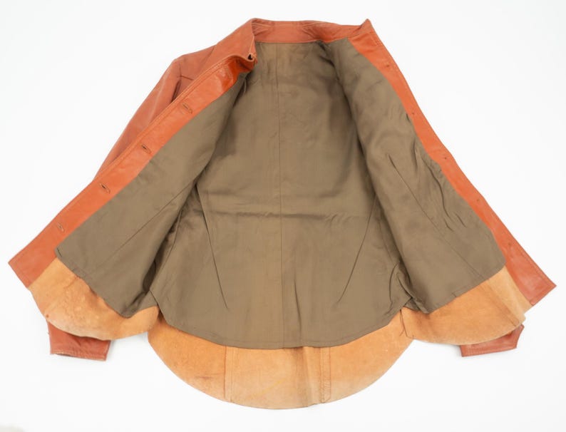May include: Open orange leather jacket with a brown interior lining. The jacket has a classic collar and button closures. The interior features a soft, suede-like texture.