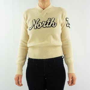 Peut inclure: Pull-over vintage crème avec un col en V et un ourlet côtelé. Le mot "North" est brodé sur la poitrine en écriture noire, avec un numéro noir sur la manche droite. Le pull est associé à un pantalon noir.