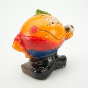 May include: A ceramic figurine of a smiling orange character wearing a red, blue, and yellow soccer uniform and holding a soccer ball. The figurine is standing on a brown base.