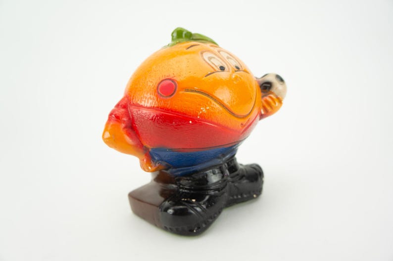 May include: A ceramic figurine of a smiling orange character wearing a red, blue, and yellow soccer uniform and holding a soccer ball. The figurine is standing on a brown base.