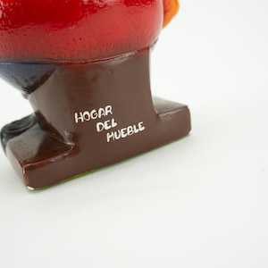 May include: A red, orange, and blue ceramic figurine of a person sitting down. The figurine is on a brown base with the text "HOGAR DEL MUEBLE" written in white.
