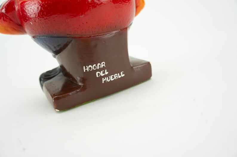 May include: A red, orange, and blue ceramic figurine of a person sitting down. The figurine is on a brown base with the text "HOGAR DEL MUEBLE" written in white.