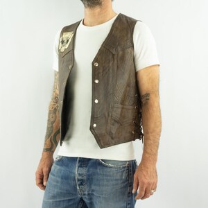 May include: Brown leather vest with a skull patch on the front. The vest has a button-up closure and side laces. The vest is worn over a white t-shirt and blue jeans.