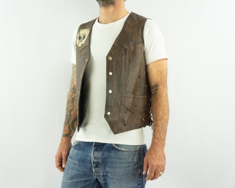 May include: Brown leather vest with a skull patch on the front. The vest has a button-up closure and side laces. The vest is worn over a white t-shirt and blue jeans.