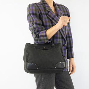 May include: A black tote bag with a flap closure and a diamond pattern. The bag has leather accents and silver-tone studs. The person is wearing a purple and green plaid blazer and grey jeans.