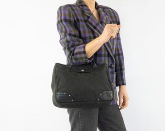 May include: A black tote bag with a flap closure and a diamond pattern. The bag has leather accents and silver-tone studs. The person is wearing a purple and green plaid blazer and gray jeans.