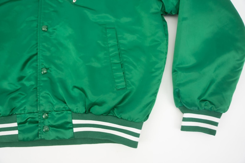 May include: Green satin bomber jacket with white and green striped cuffs and waistband. The jacket has a button closure and a pocket on the left side.