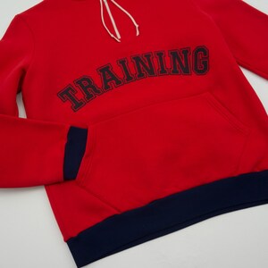 K&ouml;nnte beinhalten: Roter Kapuzenpullover mit dunkelblauem Saum und dem Wort "TRAINING" in Schwarz auf der Vorderseite gestickt.