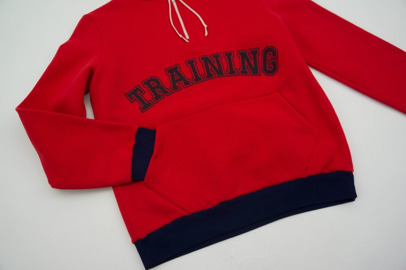 K&ouml;nnte beinhalten: Roter Kapuzenpullover mit dunkelblauem Saum und dem Wort "TRAINING" in Schwarz auf der Vorderseite gestickt.