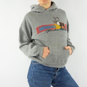 Op de afbeelding: Grijs hoodie met een grafische print van een rendier en de tekst "Anchorage Alaska".