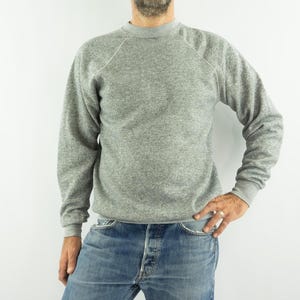 Könnte beinhalten: Ein meliertes graues Sweatshirt mit Rundhalsausschnitt und langen Ärmeln. Die Raglanärmel haben weiße Nähte. Die Bündchen und der Saum sind gerippt. Die Person auf dem Bild trägt eine blaue Jeans.