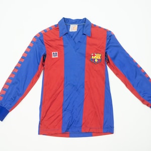 Puede incluir: Una camiseta de fútbol de manga larga con un diseño a rayas rojas y azules. La camiseta tiene cuello y mangas azules con rayas rojas. Un escudo del equipo está en el pecho y un logotipo en el lado izquierdo.