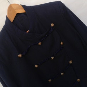 Pode incluir: Um blazer azul escuro com gola mandarim e bot&otilde;es dourados. O blazer tem uma guarni&ccedil;&atilde;o decorativa de cord&atilde;o azul escuro.