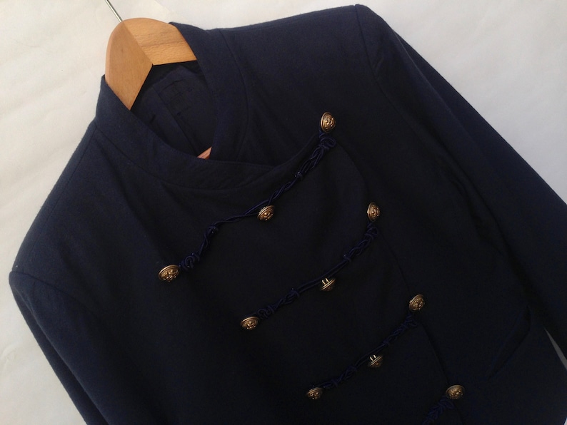Pode incluir: Um blazer azul escuro com gola mandarim e bot&otilde;es dourados. O blazer tem uma guarni&ccedil;&atilde;o decorativa de cord&atilde;o azul escuro.