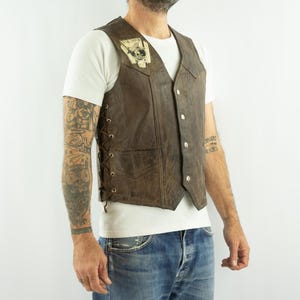 May include: Brown leather vest with a patch on the upper left chest. The vest has a button-up front, side lacing, and a pocket. The vest is worn over a white t-shirt and blue jeans.