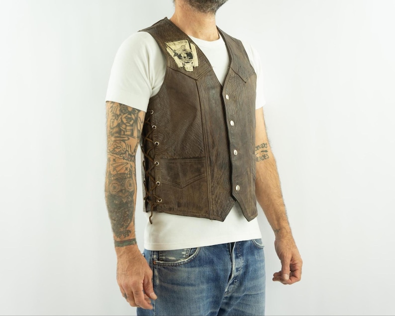 May include: Brown leather vest with a patch on the upper left chest. The vest has a button-up front, side lacing, and a pocket. The vest is worn over a white t-shirt and blue jeans.