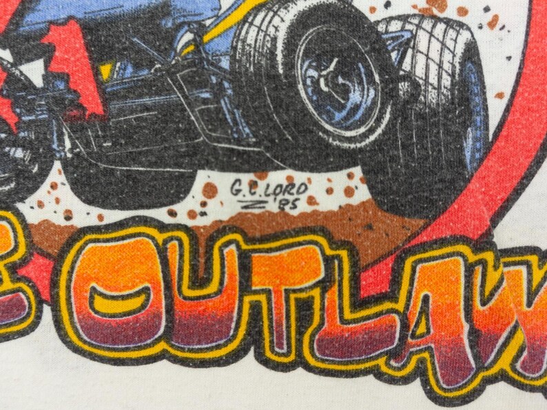 以下が含まれることがあります： 大きなタイヤが付いたレーシングカーの詳細なイラストが描かれた白いTシャツ。「OUTLAW」という大胆でカラフルなフォントでデザインされています。アーティストのサインと1995年も表示されています。