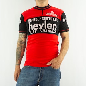 May include: A short-sleeved cycling jersey in red and black with a quarter-zip collar. The chest features white text reading MEUBEL-CENTRALE heylen VELOS PINARELLO and SPORT EN STEUN with a small wheel graphic. The sleeves have black ribbed cuffs.