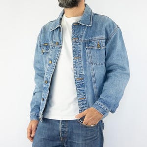 Peut inclure: Veste en jean bleu clair avec boutons et poches poitrine. La veste est portée sur un t-shirt blanc et associée à un jean bleu. Une tenue décontractée et classique.