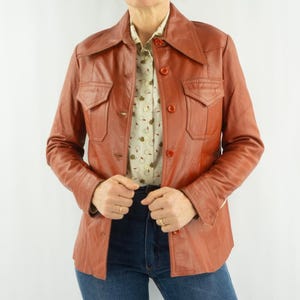 May include: A vintage rust-colored leather jacket with a collared neckline and button-down front. The jacket features two chest pockets and is worn over a patterned button-up shirt and blue jeans. The jacket has a classic, retro style.