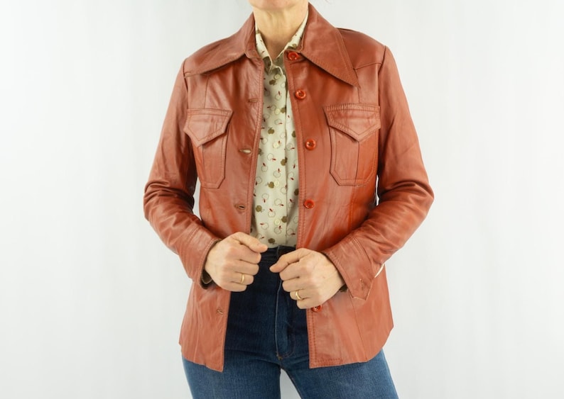 May include: A vintage rust-colored leather jacket with a collared neckline and button-down front. The jacket features two chest pockets and is worn over a patterned button-up shirt and blue jeans. The jacket has a classic, retro style.