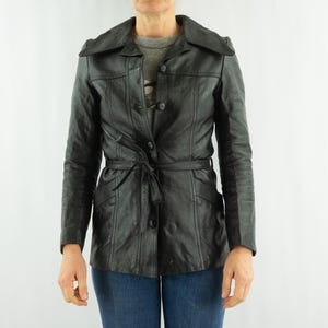 May include: Black leather jacket with a belted waist and button closure. The jacket features a notched collar and long sleeves. The jacket is approximately 28 inches long.