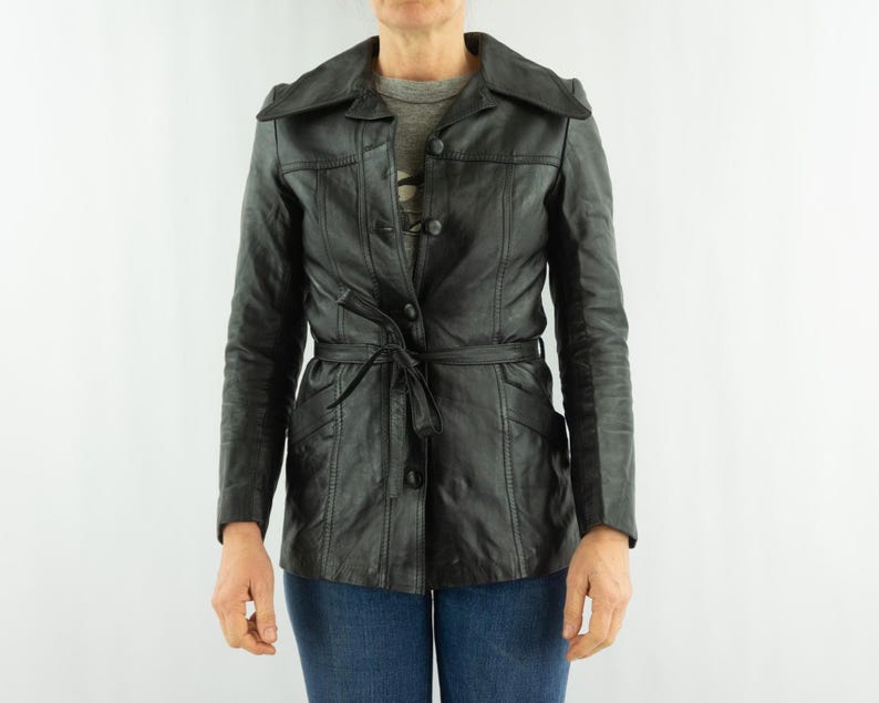 May include: Black leather jacket with a belted waist and button closure. The jacket features a notched collar and long sleeves. The jacket is approximately 28 inches long.