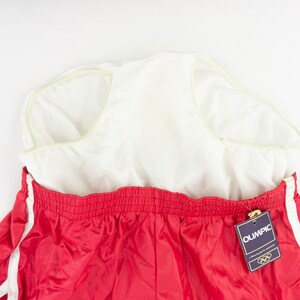 May include: Red athletic shorts with white stripes down the sides. The shorts have an elastic waistband and a white tag that says "Olympic" with the Olympic rings logo.