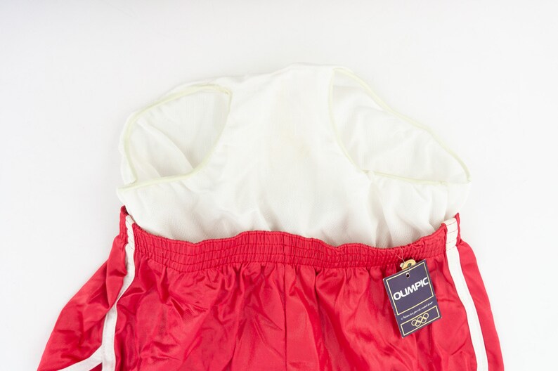 May include: Red athletic shorts with white stripes down the sides. The shorts have an elastic waistband and a white tag that says "Olympic" with the Olympic rings logo.