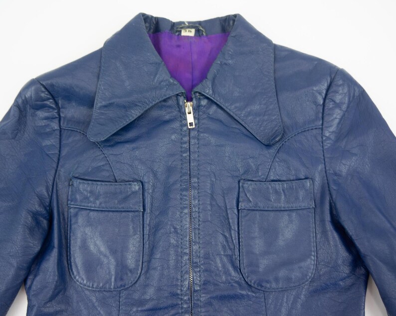 K&ouml;nnte beinhalten: Eine marineblaue Vintage-Lederjacke mit Kragen und zwei Vordertaschen. Die Jacke hat einen Rei&szlig;verschluss und ein lila Futter. Ein klassischer Stil.