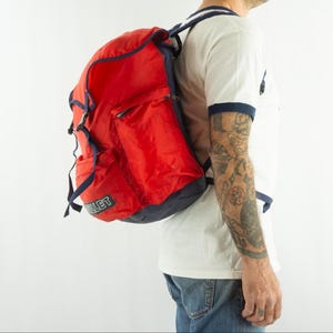 May include: A red and navy blue backpack with the word "MILLET" printed on the side. The backpack has multiple compartments and adjustable straps. The backpack is worn on the back.