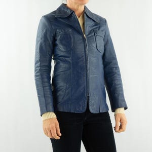 K&ouml;nnte beinhalten: Eine marineblaue Vintage-Lederjacke mit Kragen und durchgehendem Rei&szlig;verschluss. Die Jacke hat zwei Brusttaschen und wird &uuml;ber einem cremefarbenen Pullover getragen. Die Person tr&auml;gt eine schwarze Hose.