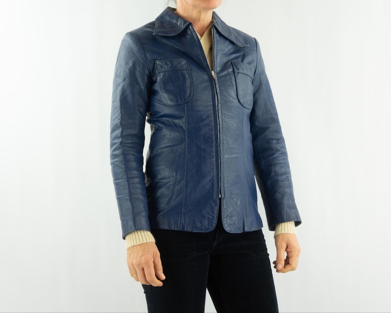 K&ouml;nnte beinhalten: Eine marineblaue Vintage-Lederjacke mit Kragen und durchgehendem Rei&szlig;verschluss. Die Jacke hat zwei Brusttaschen und wird &uuml;ber einem cremefarbenen Pullover getragen. Die Person tr&auml;gt eine schwarze Hose.