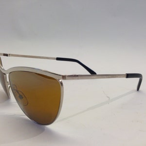 May include: Silver-toned sunglasses with brown lenses and black temple tips. The sunglasses have a cat-eye shape.