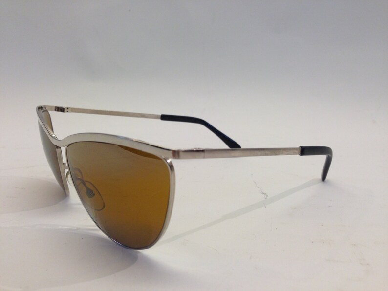 May include: Silver-toned sunglasses with brown lenses and black temple tips. The sunglasses have a cat-eye shape.