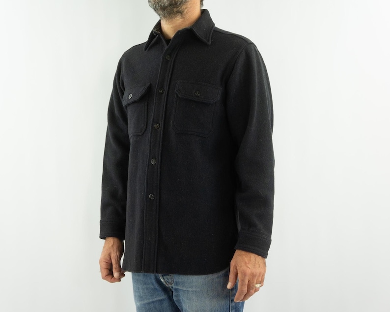 May include: A dark charcoal button-down shirt with two chest pockets. The shirt has a classic collar and is made of a textured fabric. The sleeves are long, and the shirt is worn with blue jeans. A casual, everyday garment.