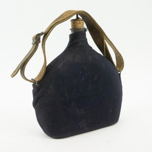 Puede incluir: Una cantimplora de color azul marino oscuro cubierta con una funda de tela, que presenta una correa de transporte de cuero marrón claro con una hebilla de metal y un tapón de corcho en la parte superior. Tiene una forma redondeada y plana.