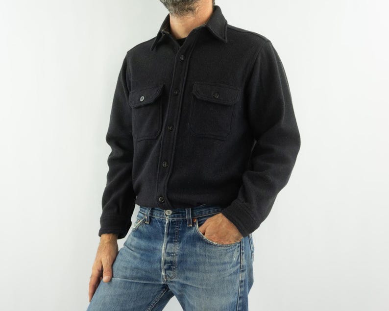 May include: A dark gray button-down shirt with two chest pockets. The shirt has a collar and long sleeves. The person in the image is wearing blue jeans. The shirt is made of a thick, textured fabric.